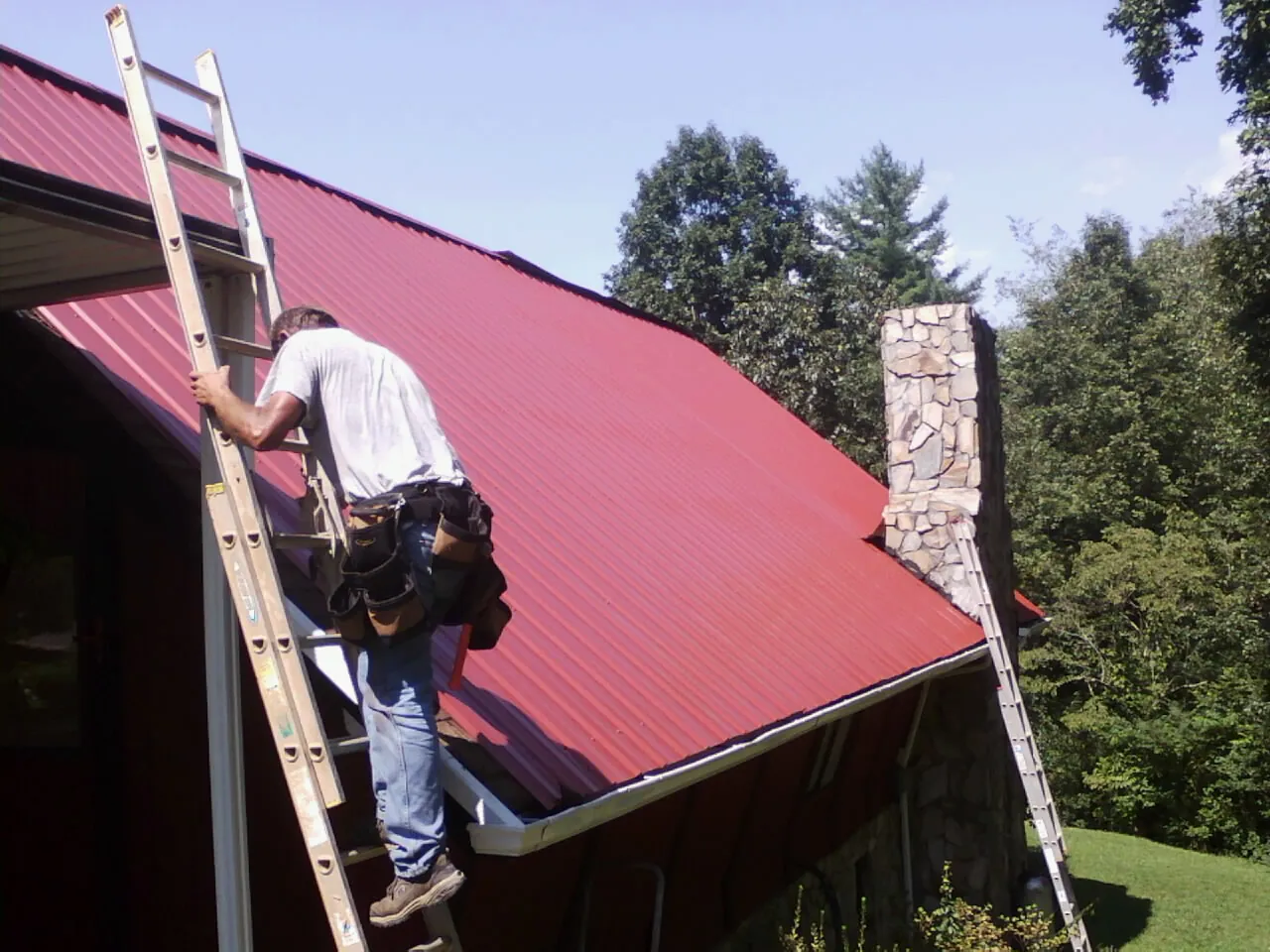 Roofer on a ladder beside a red metal roof and stone chimney — Soffit & Fascia Repair by Roofing Brothers in Barnum